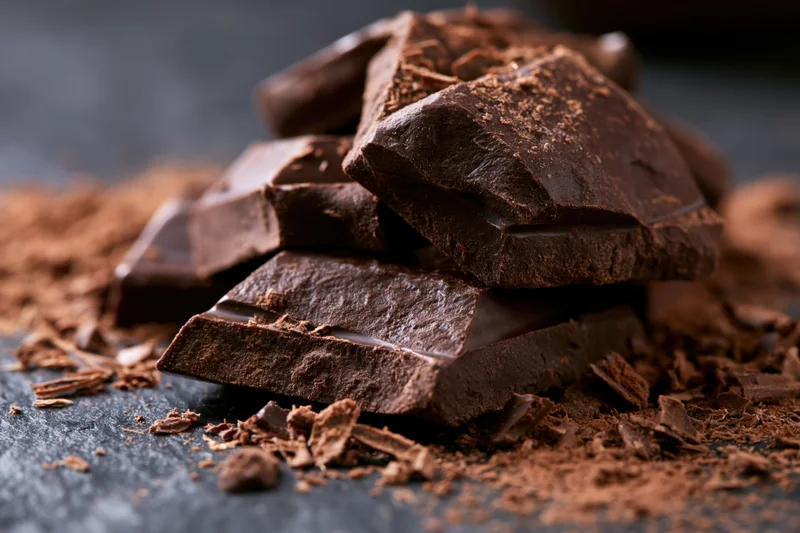 Broken dark chocolate chunks piled haphazardly on a dark surface with cocoa powder scattered around