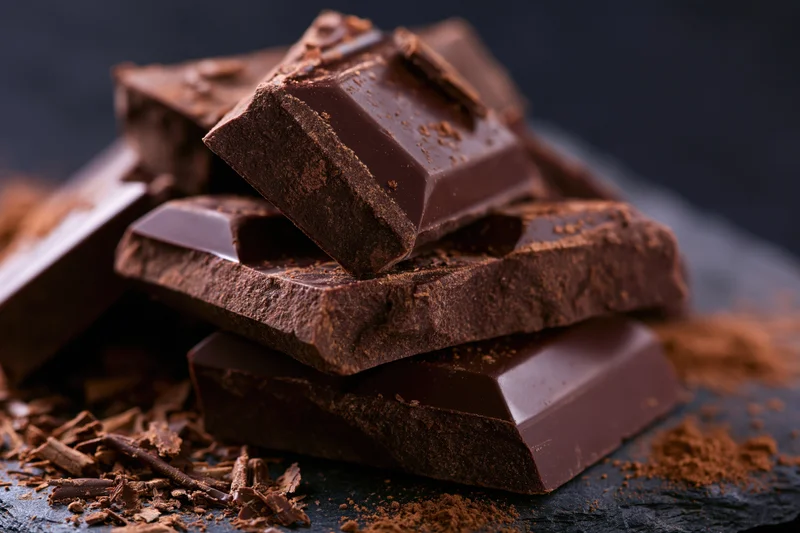 Broken dark chocolate chunks piled haphazardly on a slate surface with cocoa powder and shavings scattered around