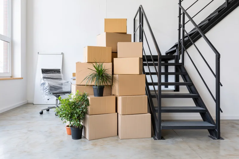 Stacked cardboard moving boxes and potted plants beside a metal staircase in a modern loft interior