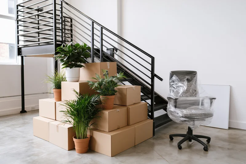 Stacked cardboard moving boxes and potted plants clustered near a metal staircase in a modern loft interior