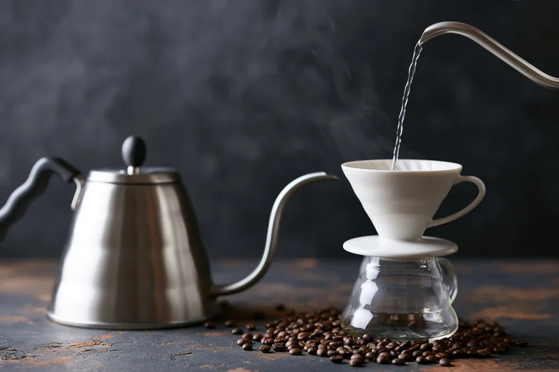 Hot water poured from a gooseneck kettle into a ceramic filter over a glass carafe surrounded by coffee beans on a dark rustic surface