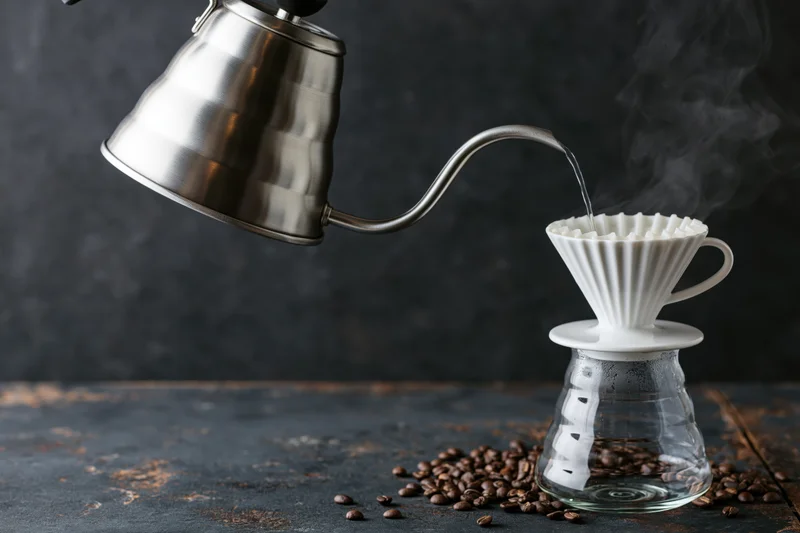 Stainless steel gooseneck kettle pouring hot water over coffee grounds in a white ceramic filter on a glass carafe, with scattered beans on a dark rustic surface
