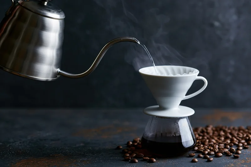 Stainless steel gooseneck kettle pouring hot water into a white ceramic filter over a glass carafe surrounded by coffee beans on a dark surface