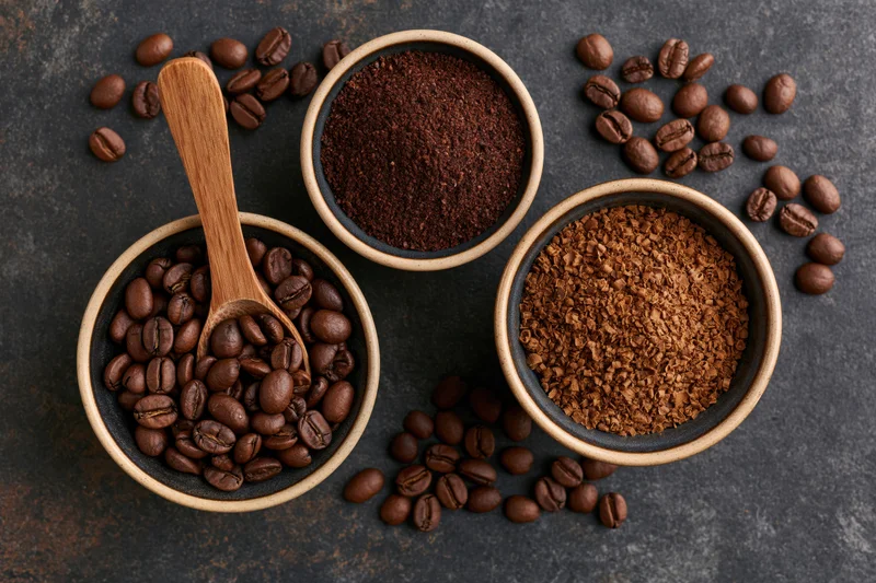 Three bowls with coffee beans, ground coffee and instant coffee arranged on dark stone surface with wooden spoon