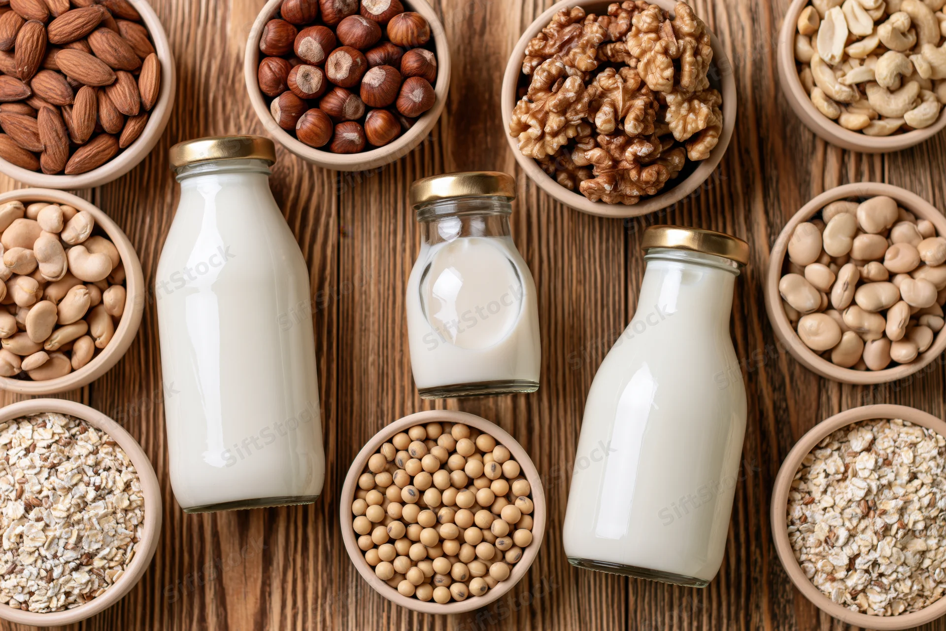Three glass bottles of white plant-based milk arranged with bowls of nuts, seeds and grains on a wooden table in a flat lay top view