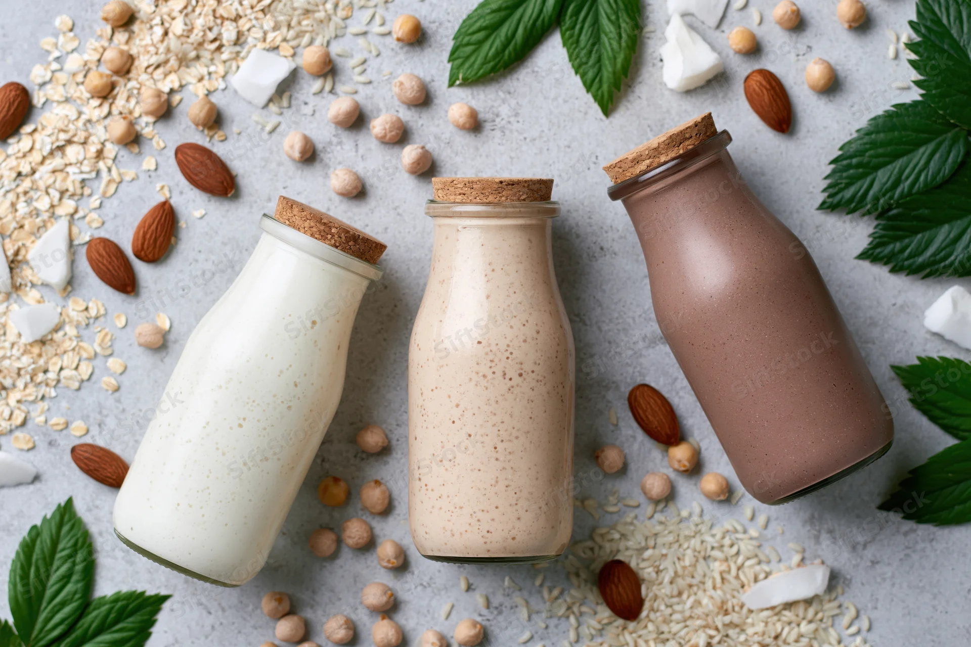 Three glass bottles of plant-based milk arranged with nuts, seeds, and leaves on a gray surface