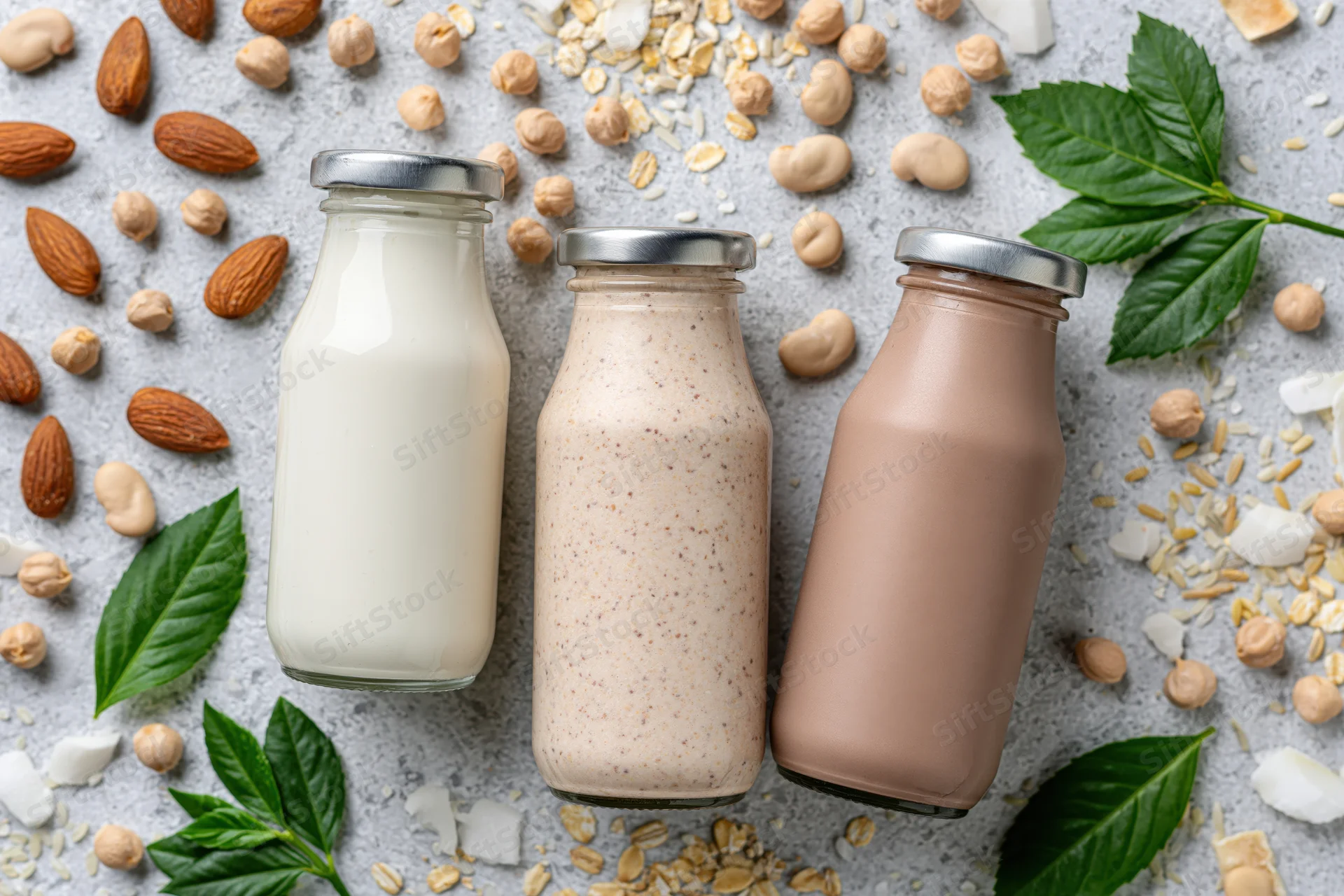Three glass bottles of plant-based milk arranged on a gray surface with scattered nuts, seeds, and leaves