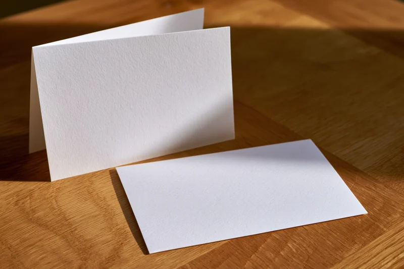 Two blank white folded greeting cards on a warm oak wooden table with sunlight and shadows