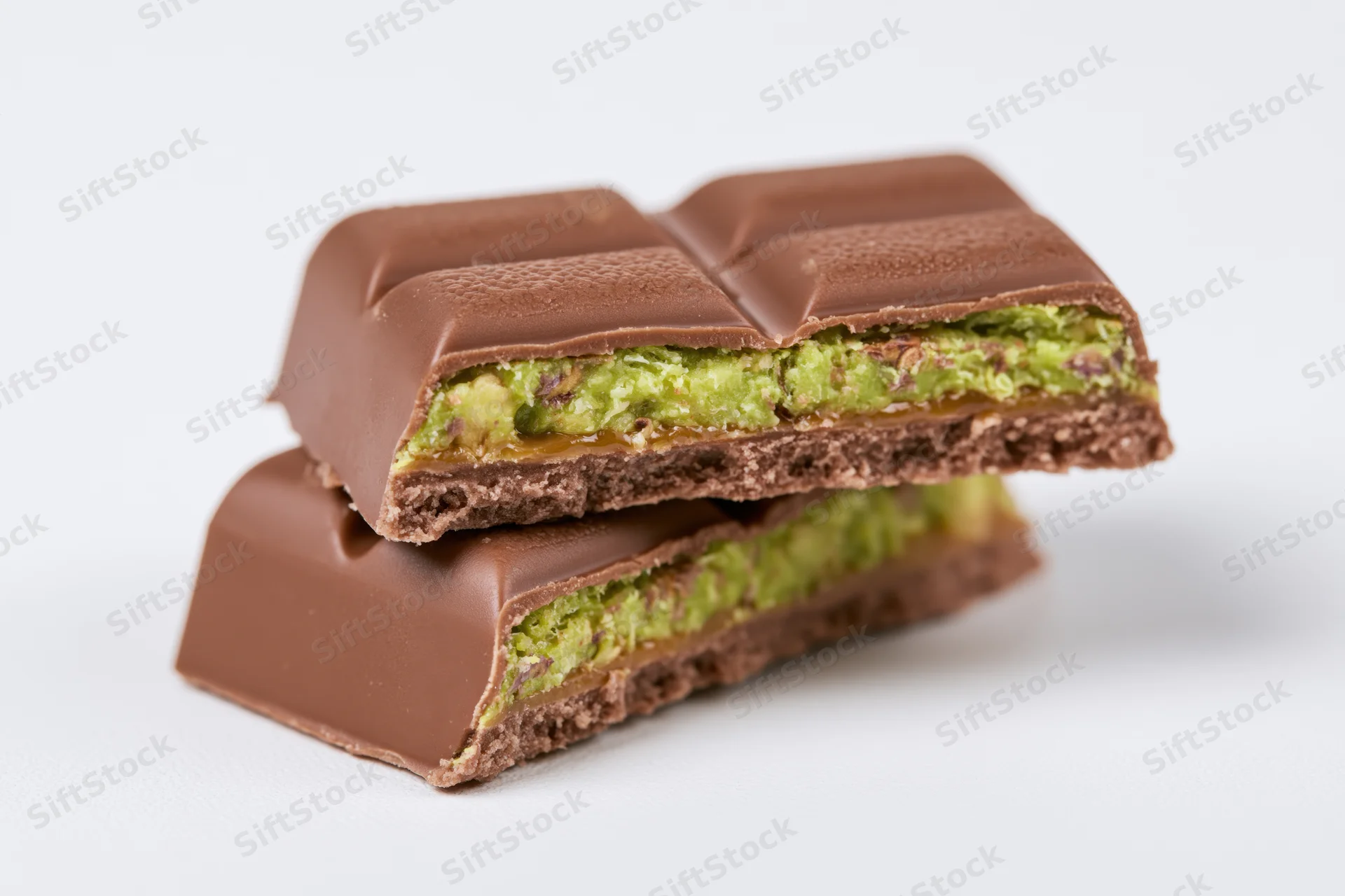 Two broken milk chocolate bar pieces stacked on white background showing green pistachio and caramel filling