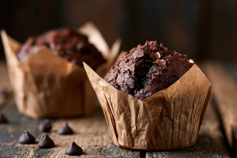 Two dark chocolate muffins in crinkled paper wrappers on wood