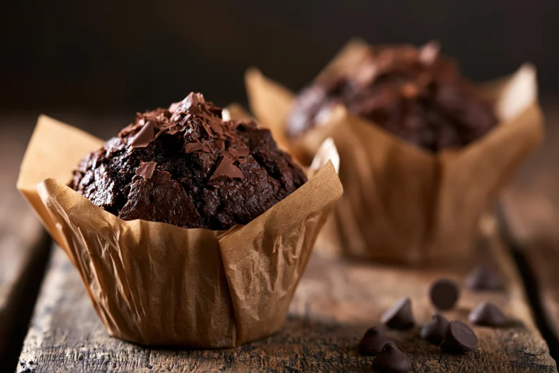 Two dark chocolate muffins in crinkled brown paper wrappers on a wooden surface with scattered chocolate chips