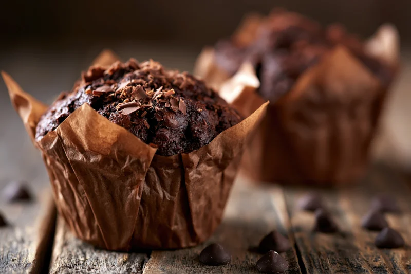 Two dark chocolate muffins in brown paper wrappers on wood