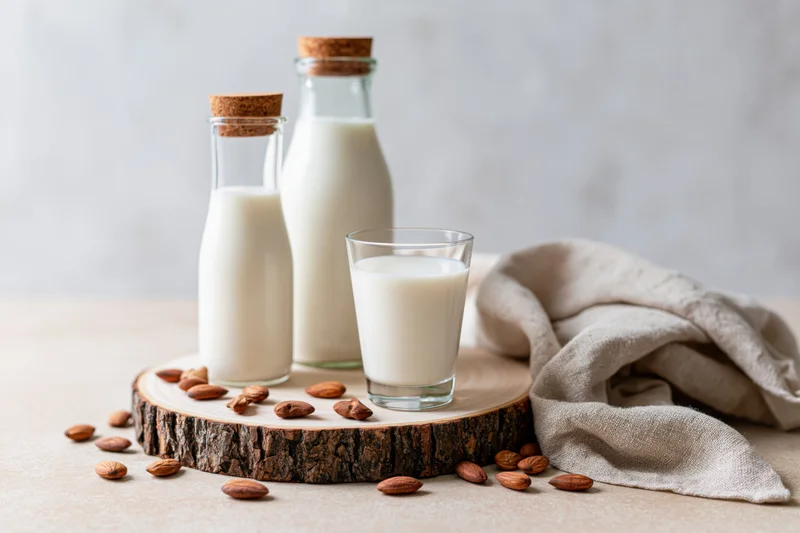 Almond milk in glass bottles and a glass on a wooden slice with scattered almonds and a linen cloth on a light surface