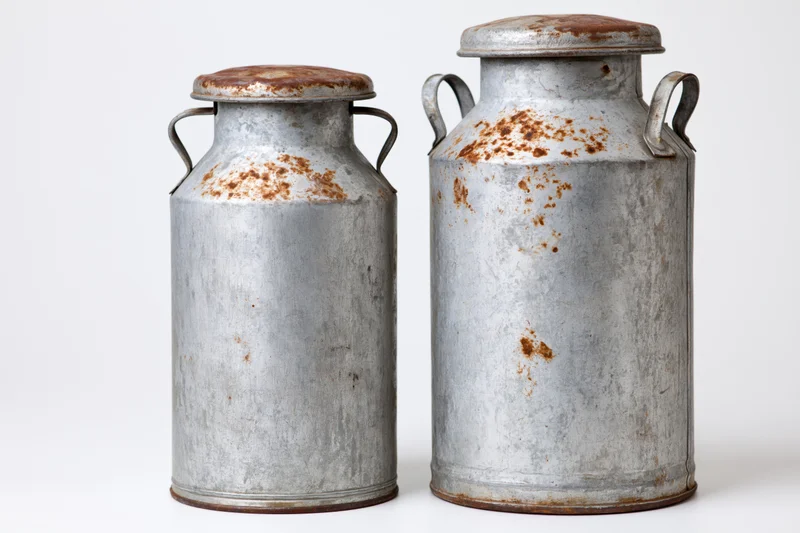 Two old metal milk cans of different heights standing side by side on white background