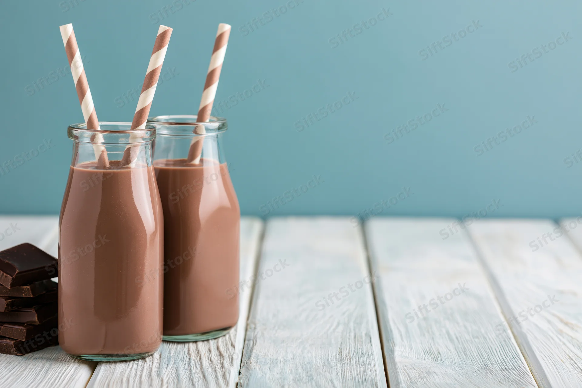 Two glass bottles of chocolate milk with striped straws on a white wooden table