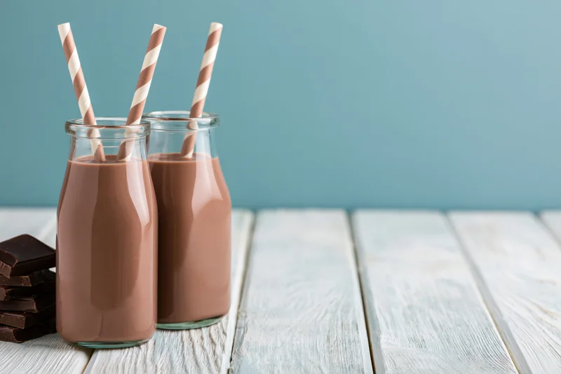Two glass bottles of chocolate milk with striped straws on a white wooden table