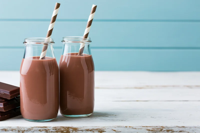 Two glass bottles filled with chocolate milk on a white wooden table with striped straws and chocolate bars beside them