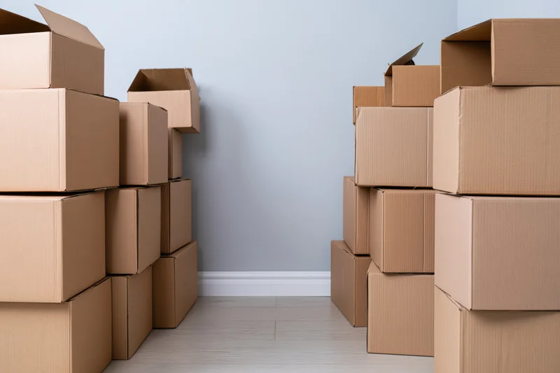 Stacks of brown cardboard moving boxes arranged on both sides of a room with light wood flooring and pale blue walls