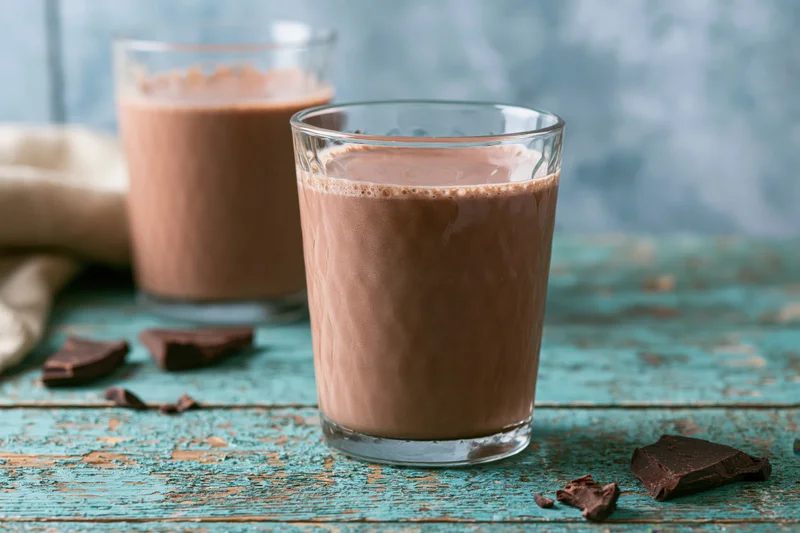 Two glasses of creamy chocolate milk on a weathered turquoise wooden table with chocolate chunks scattered around