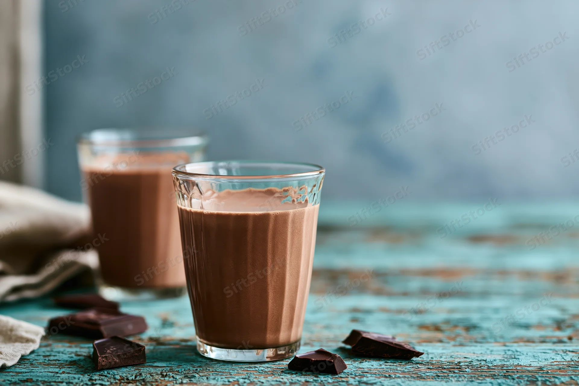 Two textured glass tumblers filled with creamy chocolate milk on a rustic blue wooden table with chocolate pieces scattered around
