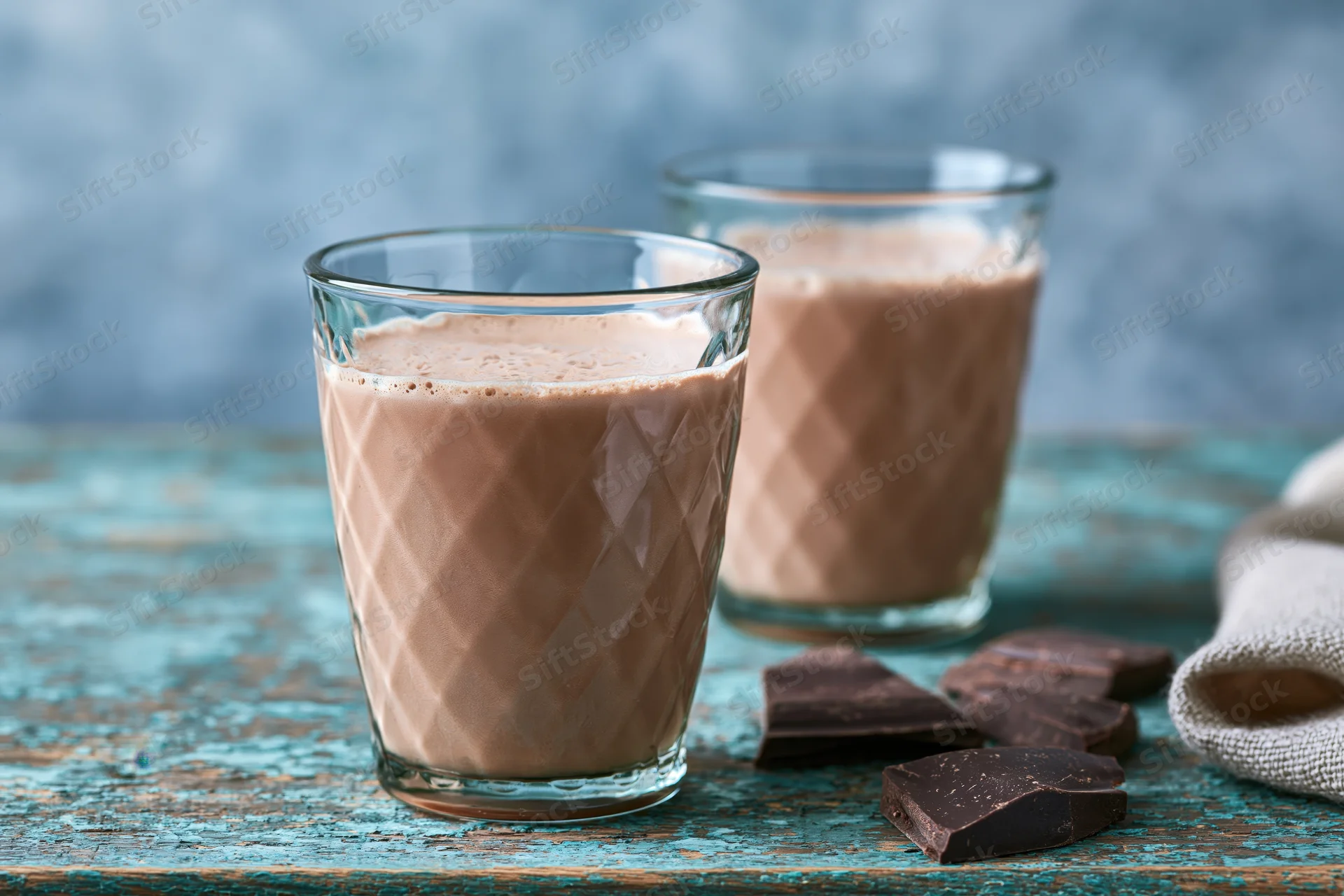 Two textured glass tumblers filled with creamy chocolate milk on a rustic wooden table with chocolate pieces beside