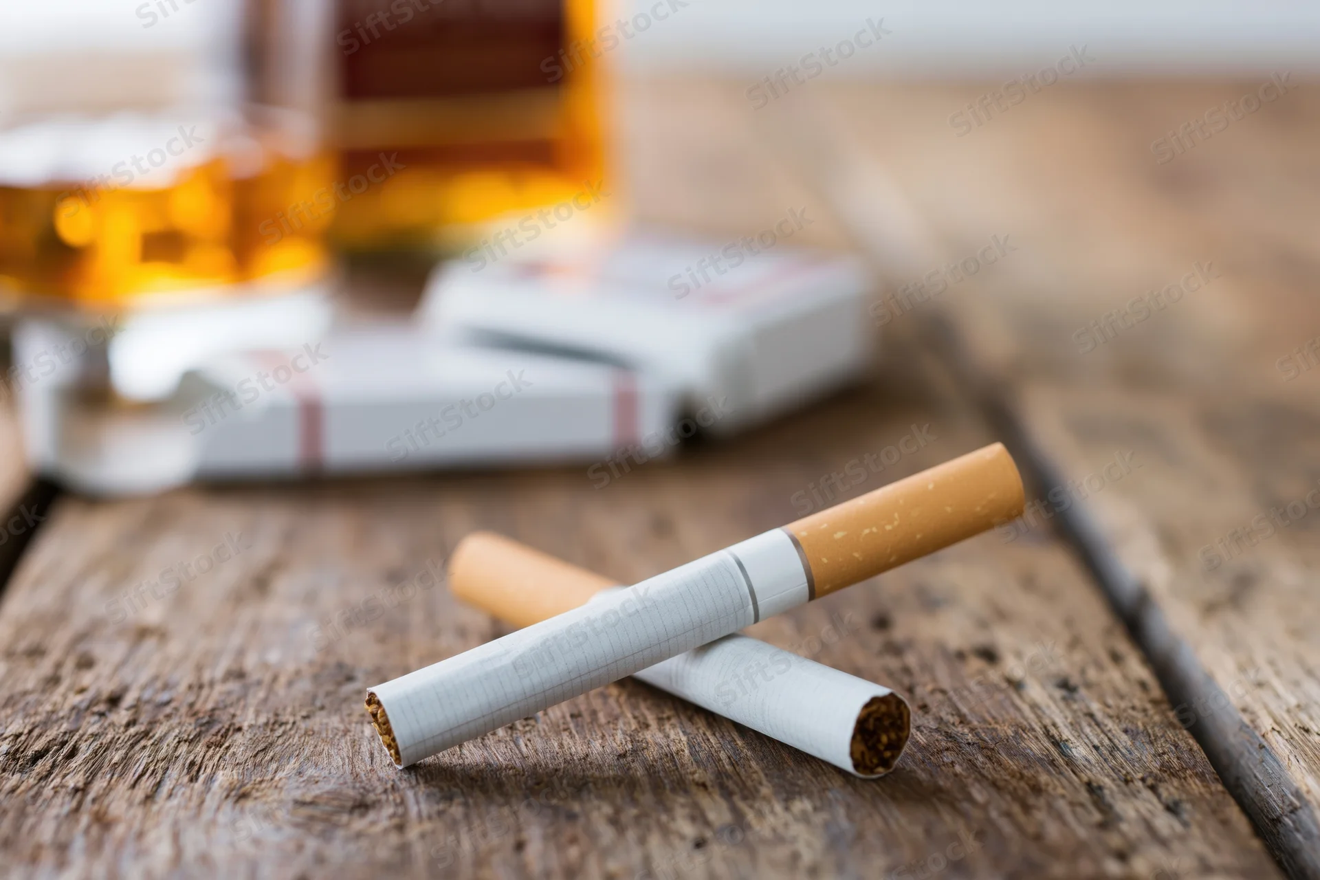 Two unlit cigarettes crossed on a worn wooden table with whiskey bottle and pack in background