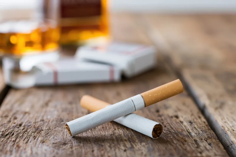 Two unlit cigarettes crossed on a worn wooden table with whiskey bottle and pack in background