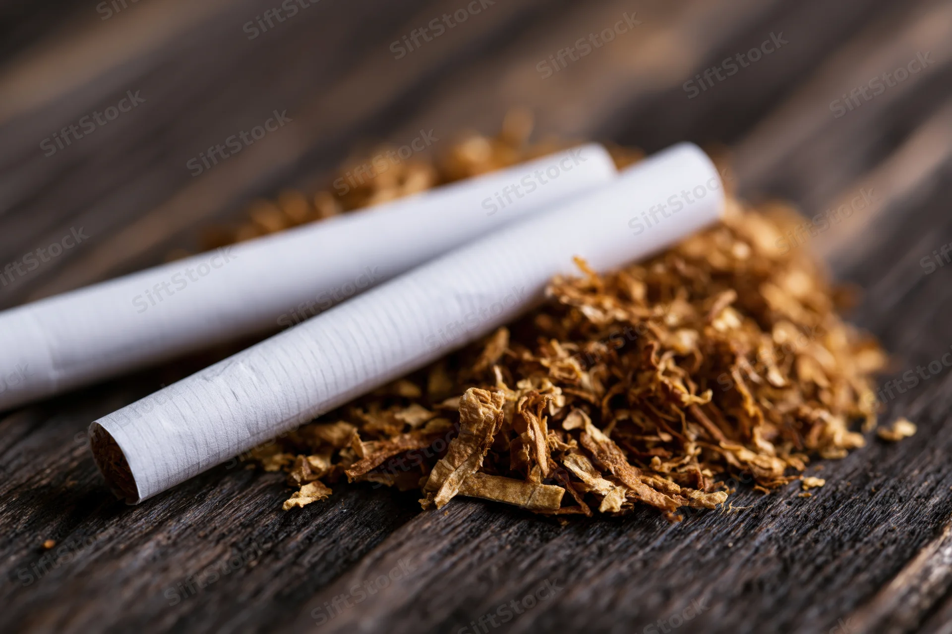 Two unlit cigarettes resting on a pile of dried tobacco leaves on a dark wooden surface