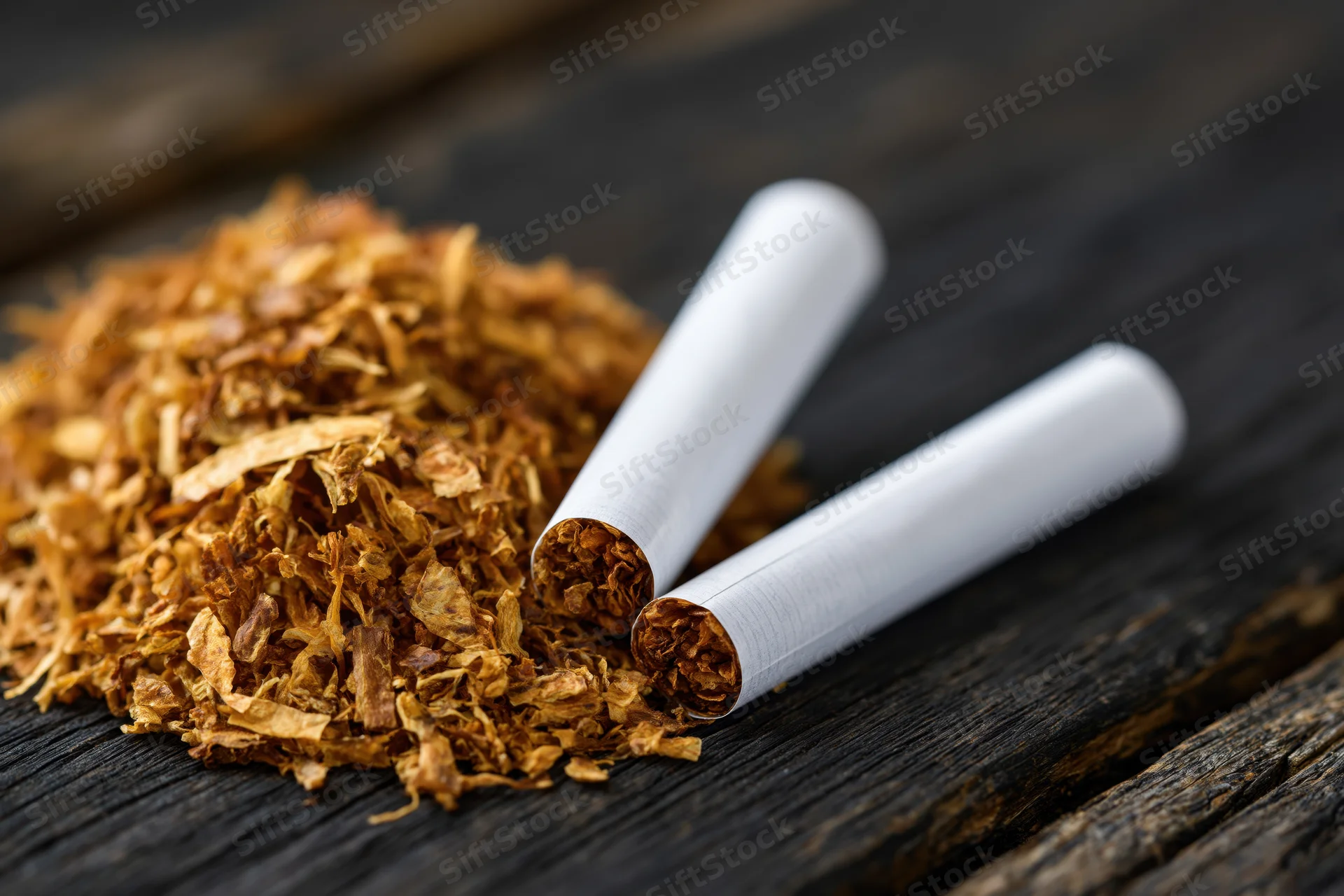 Two unlit cigarettes resting against a pile of dried loose tobacco on dark wooden surface