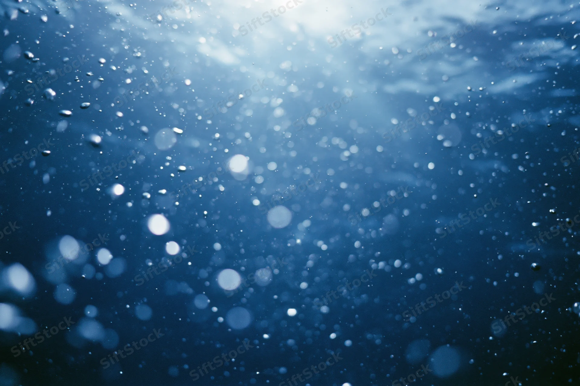 Underwater view looking up toward sunlit surface with rising air bubbles in deep blue ocean