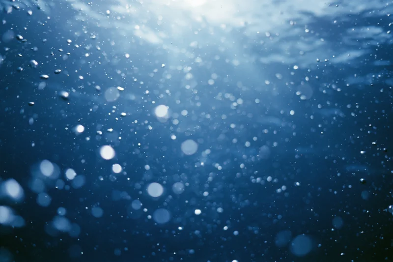 Underwater view looking up toward sunlit surface with rising air bubbles in deep blue ocean
