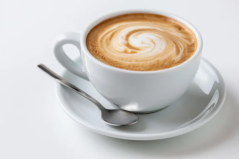 White ceramic cappuccino cup with latte art swirl on matching saucer and spoon, isolated on white background