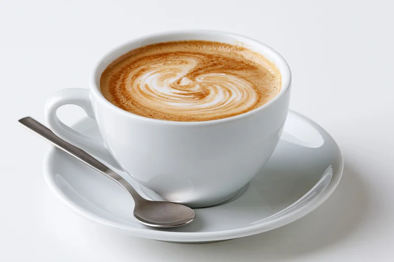 White ceramic cappuccino cup with latte art swirl on matching saucer and spoon, isolated on white background