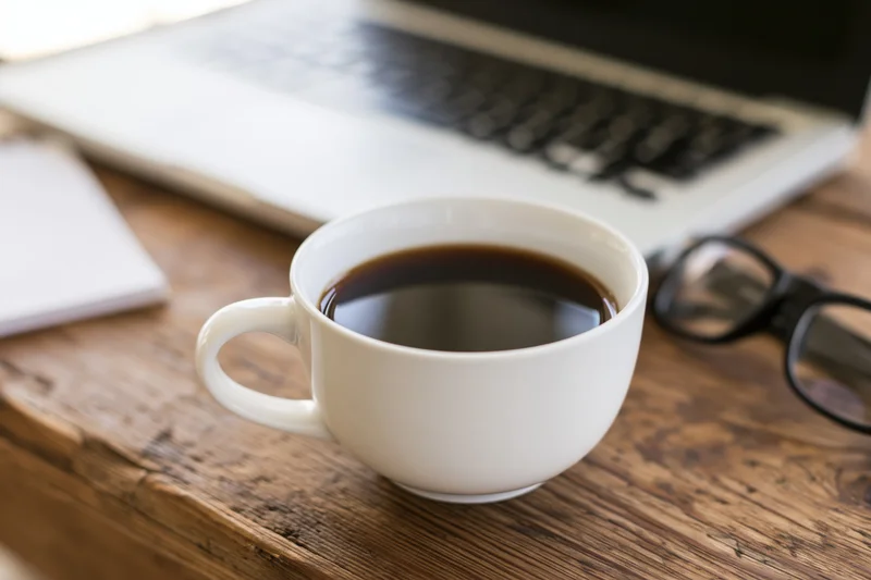White ceramic coffee cup filled with black coffee on wooden desk beside laptop and glasses