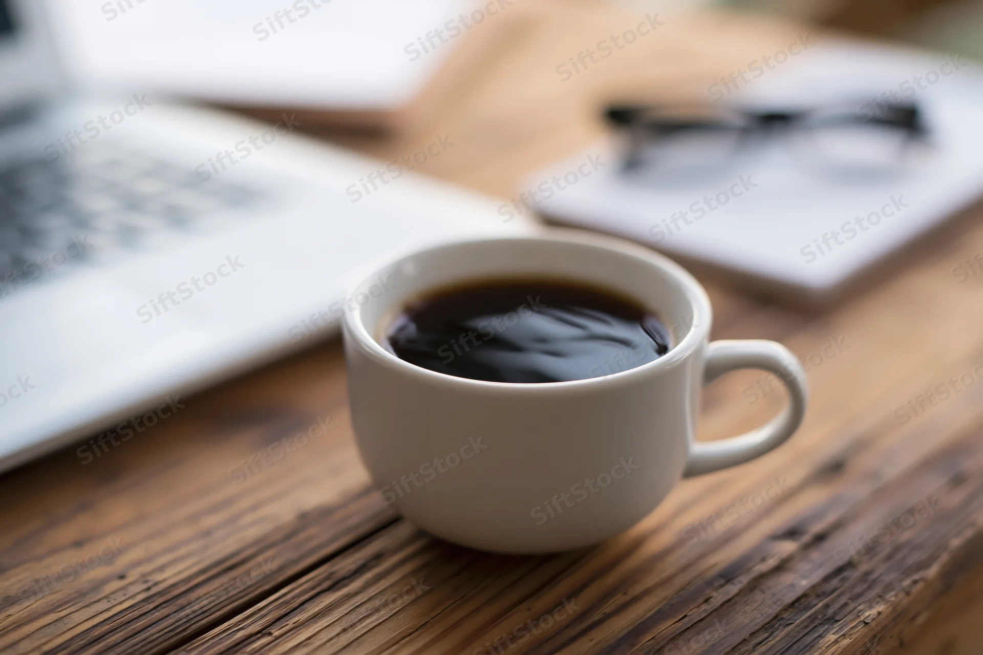 White ceramic coffee cup filled with black coffee on wooden desk beside laptop and glasses