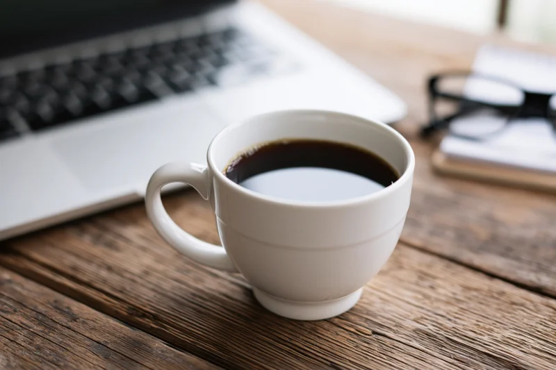 White ceramic coffee cup filled with black coffee on wooden desk beside laptop and glasses