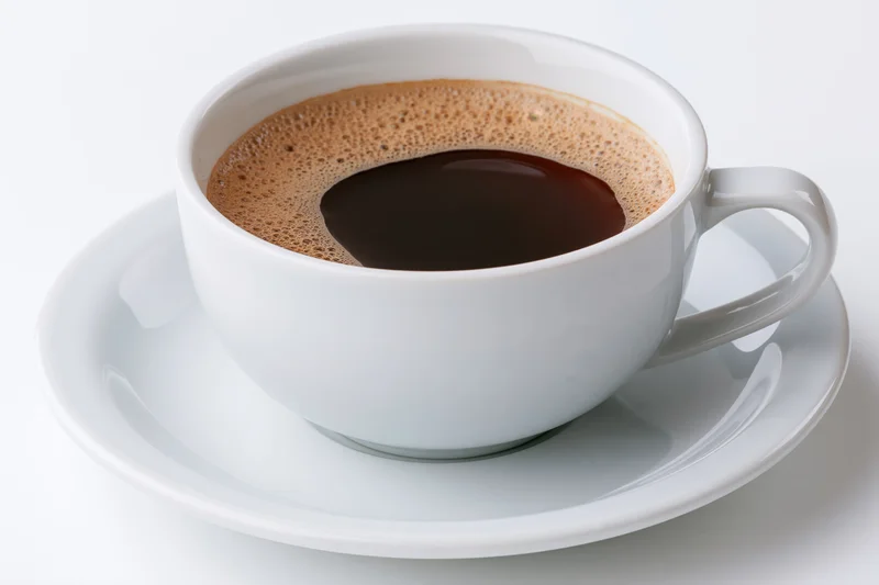 White ceramic coffee cup filled with black coffee topped with foam on a saucer against a white background