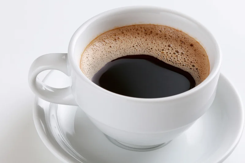 White ceramic coffee cup filled with black coffee topped with foam on a saucer against a white background