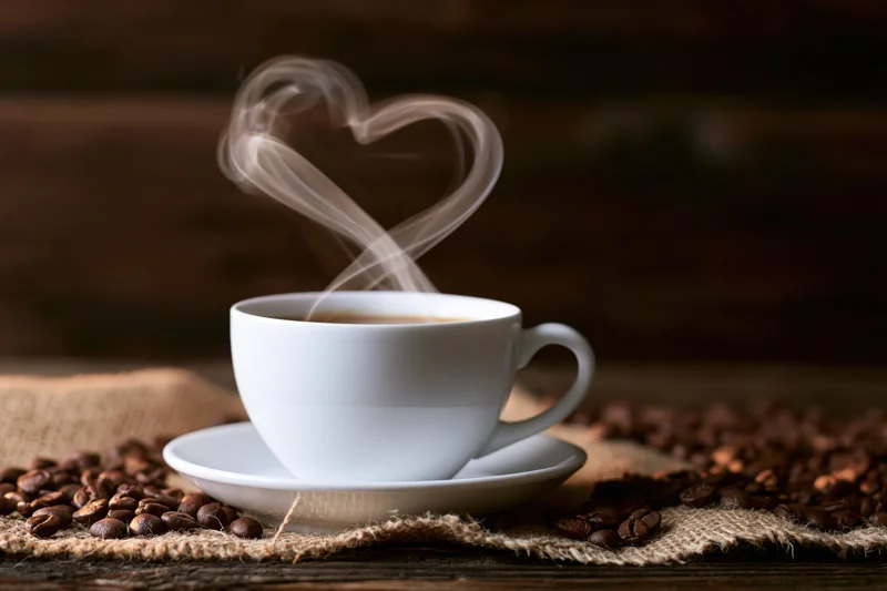 White ceramic coffee cup on saucer with heart-shaped steam rising, surrounded by coffee beans on burlap over wooden table