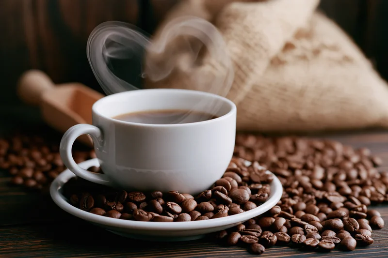 White ceramic coffee cup with heart-shaped steam on saucer surrounded by roasted beans on dark wooden table