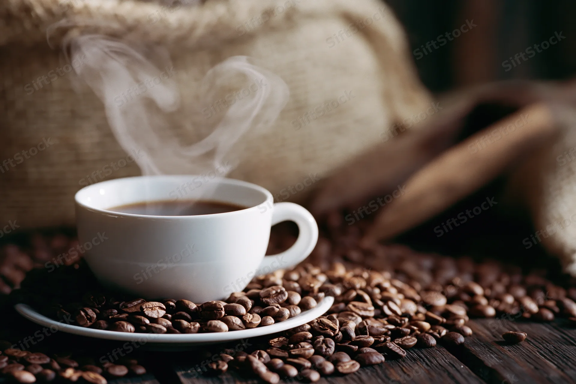 Steaming white ceramic coffee cup on saucer surrounded by scattered roasted coffee beans on dark wooden table