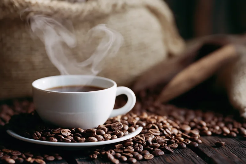 Steaming white ceramic coffee cup on saucer surrounded by scattered roasted coffee beans on dark wooden table