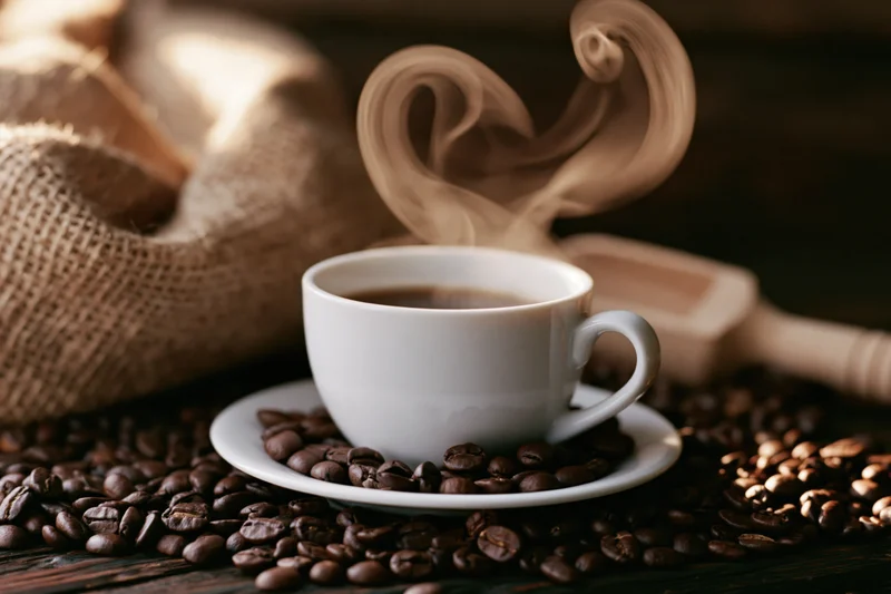 White ceramic coffee cup with heart-shaped steam on saucer surrounded by roasted coffee beans on dark wooden table