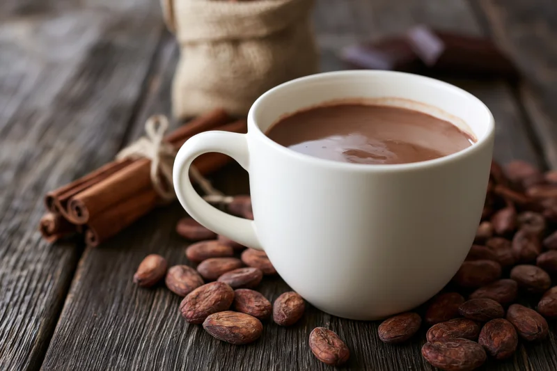 White ceramic cup filled with hot chocolate surrounded by cocoa beans and cinnamon sticks on a rustic wooden table
