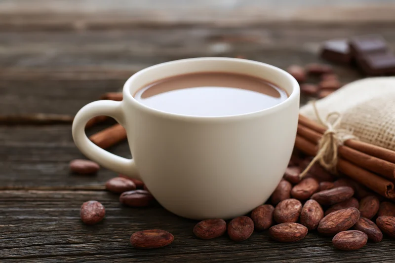 White ceramic cup filled with hot chocolate surrounded by cocoa beans and cinnamon sticks on a wooden table