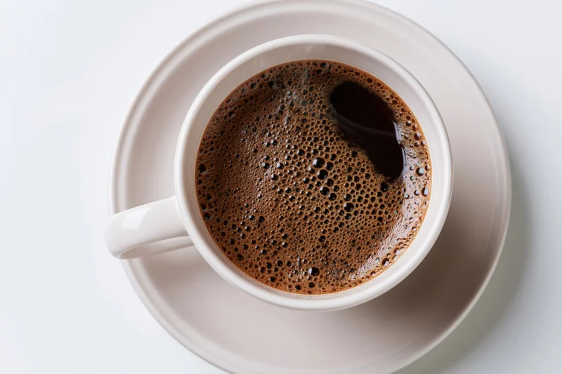 White ceramic cup of black coffee with foam on matching saucer viewed from above on plain surface