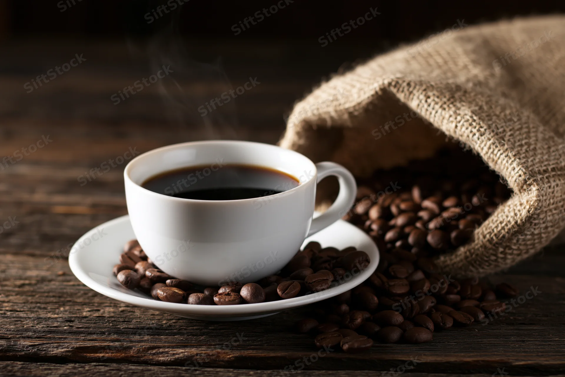 Steaming black coffee in a white ceramic cup on a saucer surrounded by roasted coffee beans and a burlap sack on a dark wooden table