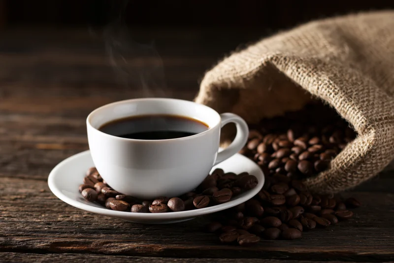 Steaming black coffee in a white ceramic cup on a saucer surrounded by roasted coffee beans and a burlap sack on a dark wooden table