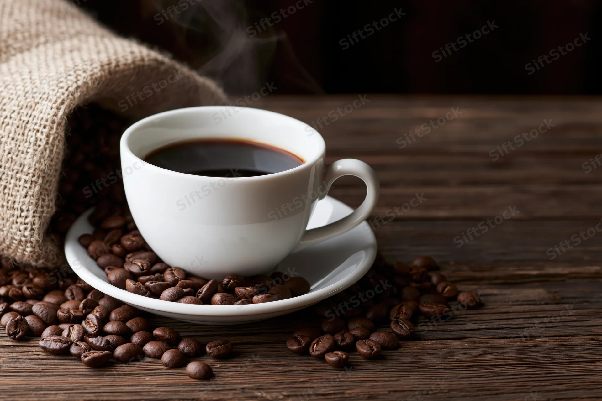 Steaming black coffee in a white ceramic cup on a saucer surrounded by roasted coffee beans on a wooden table