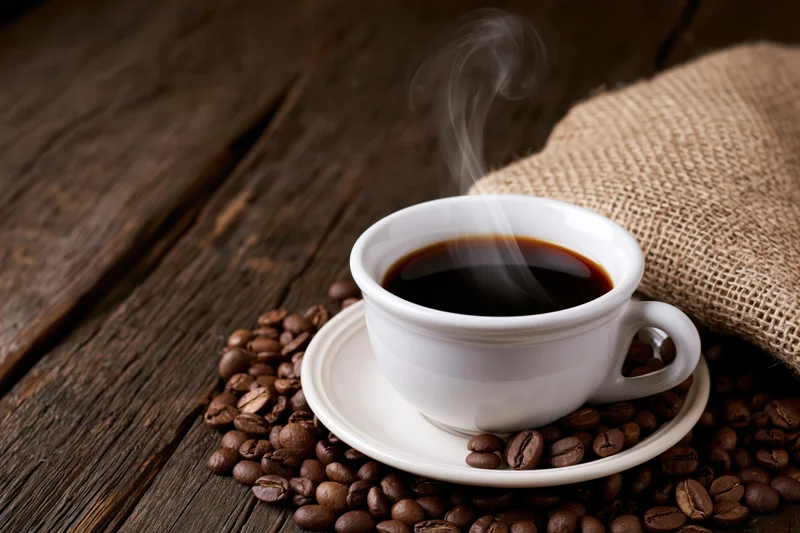 Steaming black coffee in a white ceramic cup on a saucer surrounded by roasted beans on a wooden table with burlap sack